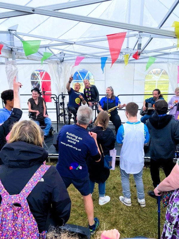 A group of people are dancing inside a white tent adorned with colourful bunting. They are facing a stage of people playing different instruments and singing.