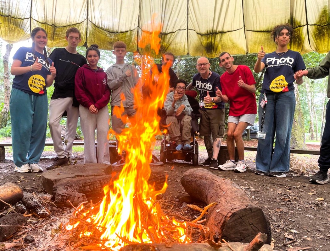 A group of adults and children are grouped together underneath a large canopy, standing behind a fire. They are smiling and giving thumbs up.