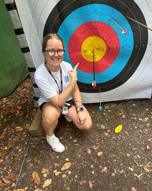 A young adult is kneeling in front of an archery board, smiling and pointing at an arrow in the centre.