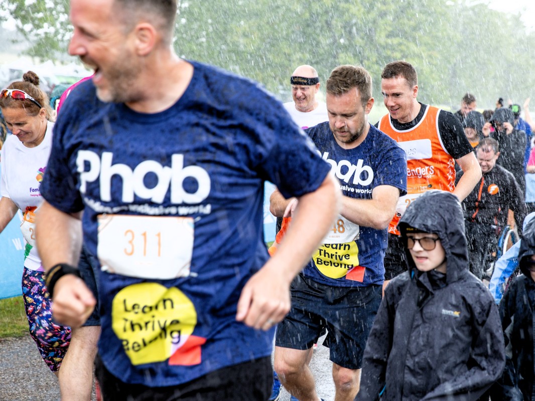 A group of adults and children, some wearing blue t-shirts with Phab on, are running together through torential rain. You can see other people running behind them.