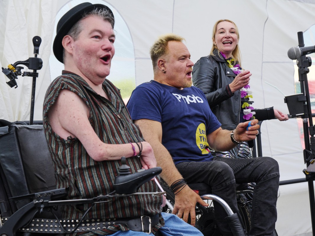 Three people,  two using a wheelchair each, one standing, are singing together side by side.