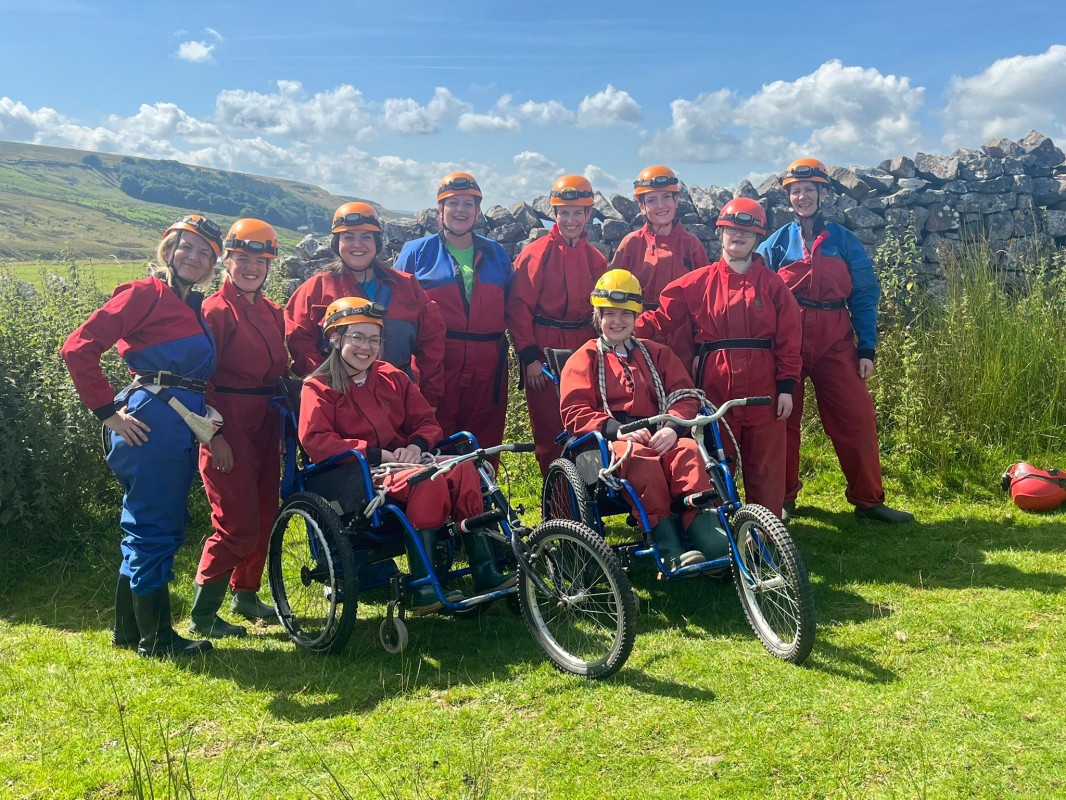 10 adults and children wearing red and blue caving cuits and orange helmets are grouped together outside, with a view of rolling hills and blue sky behind them. They are all smiling, and two people are seated in three wheeled wheelchairs.