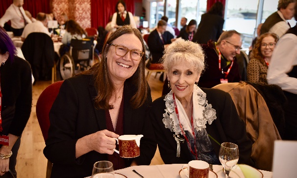 Two people are sat side by side smiling at a table adorned with mugs and wine glasses. One is raising a mug, the other is Anita Dobson. Behind them are groups of people sat at other tables.
