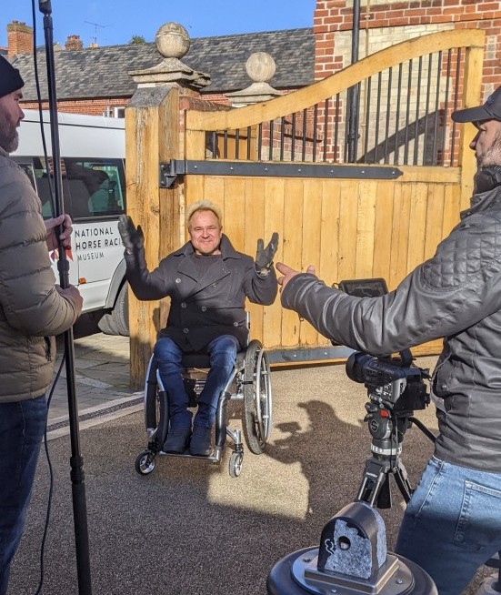 Mik, a white wheelchair user with dyed blonde hair, presents to camera while two men on the corners of the photograph film him, one suing a camera the other holding a boom mic