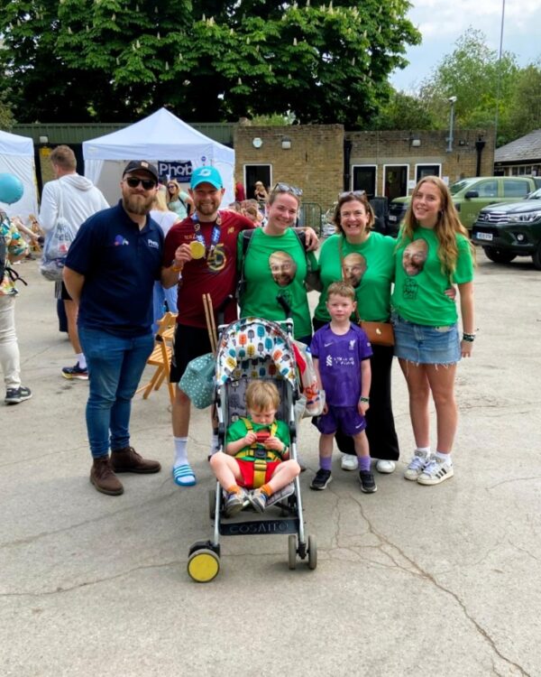 Five adults and two children, one sat in a buggy, are arm in arm posing together. Three people are wearing green t-shirts with a face on them, and one is holding a medal.