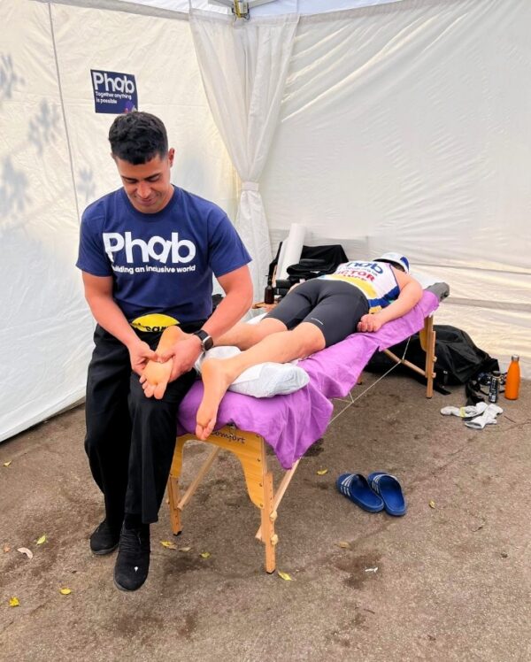 A person wearing a blue top reading "Phab" is sat on the end of a table massaging a persons foot.