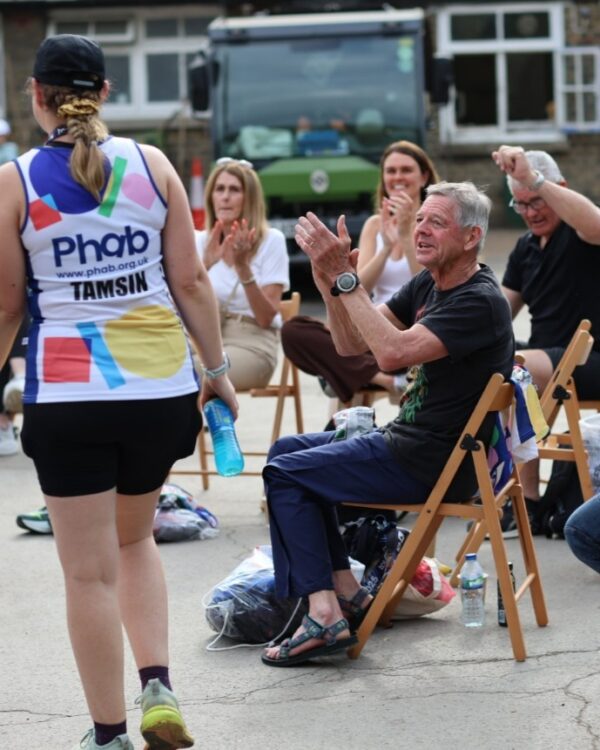 A person wearing a vest reading "Phab" is walking past a group of people sat down and clapping.
