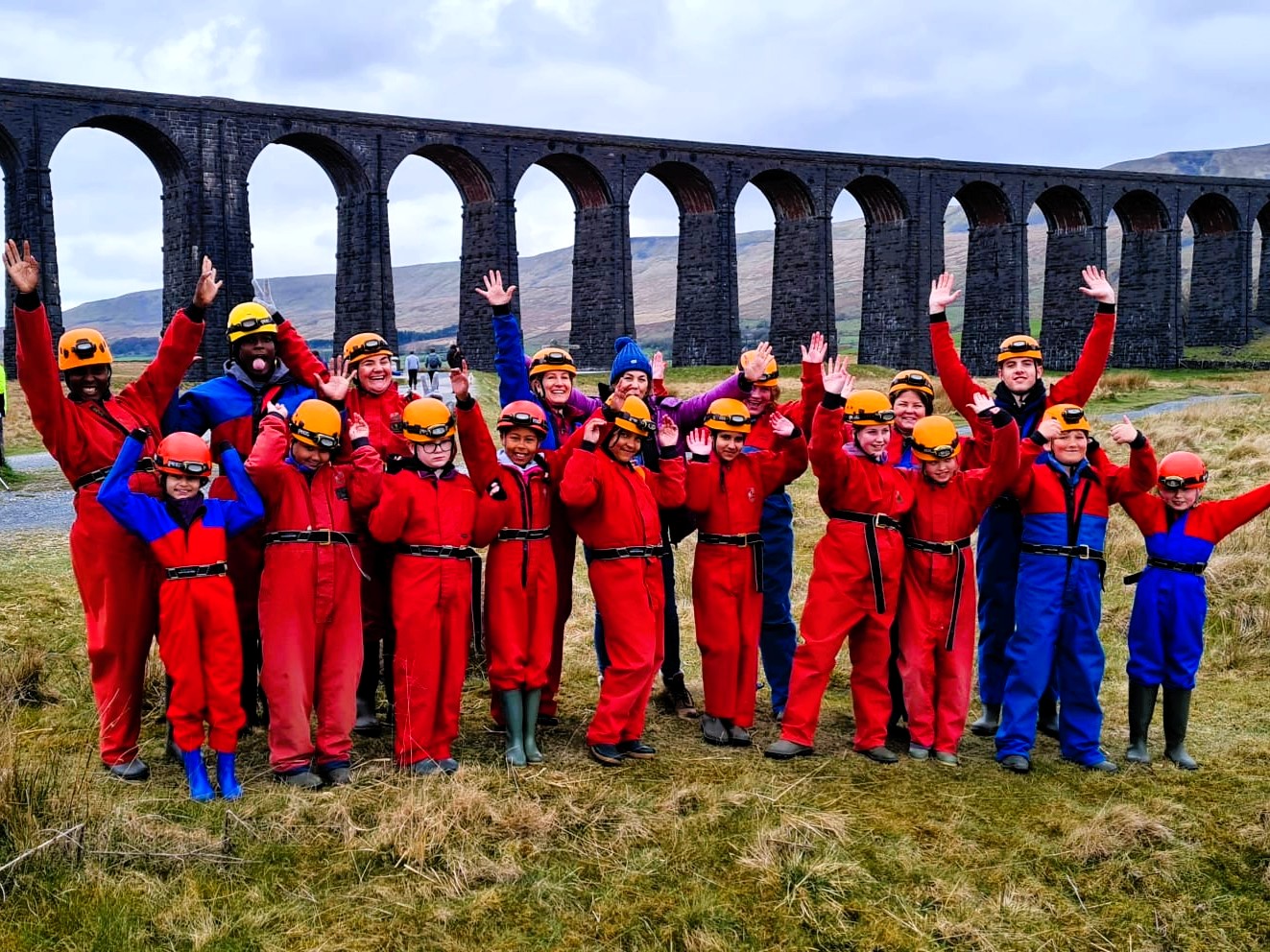 A group of children and adults wearing red or blue caving suits, are stood together in front of a very large stone bridge, cheering and waving.