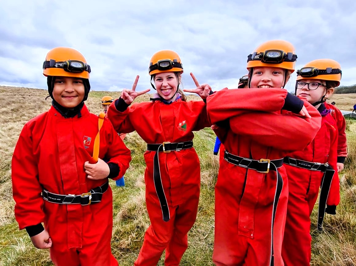 Four children wearing red caving suits and orange helmets are smiling or making peace signs together.