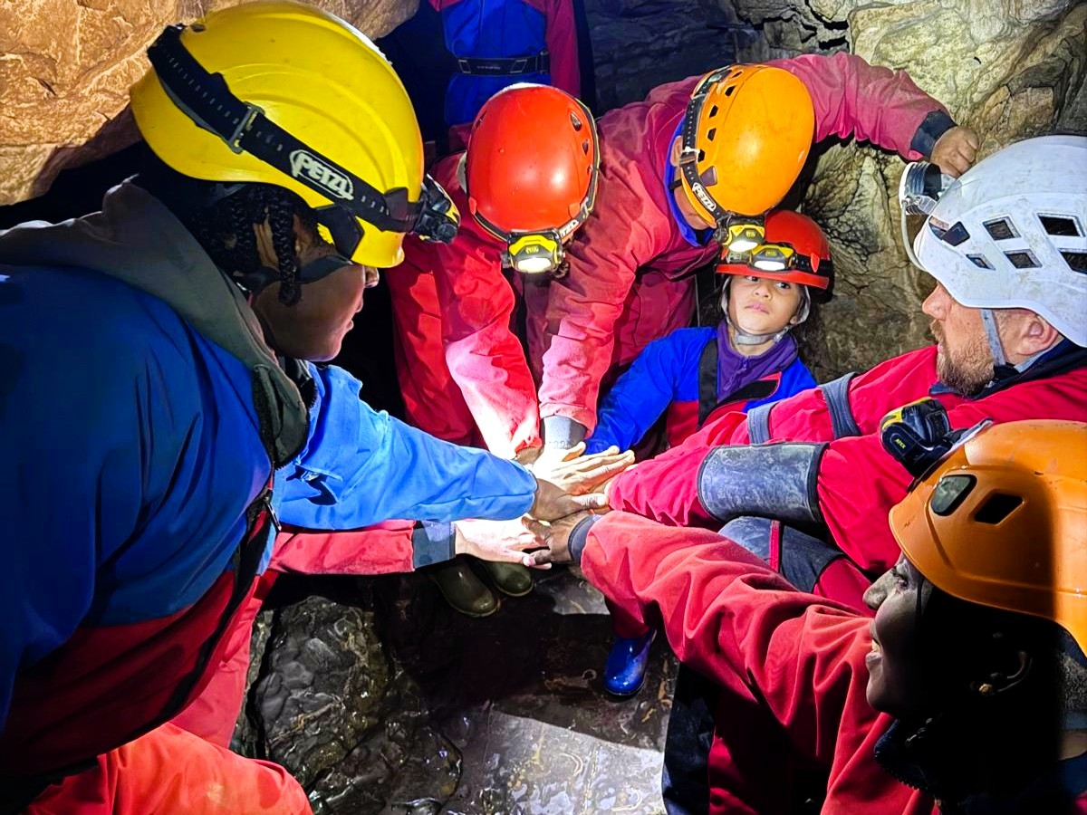 A group of people and children are wearing red or blue caving suits and helmets, putting their hands on top of each other.
