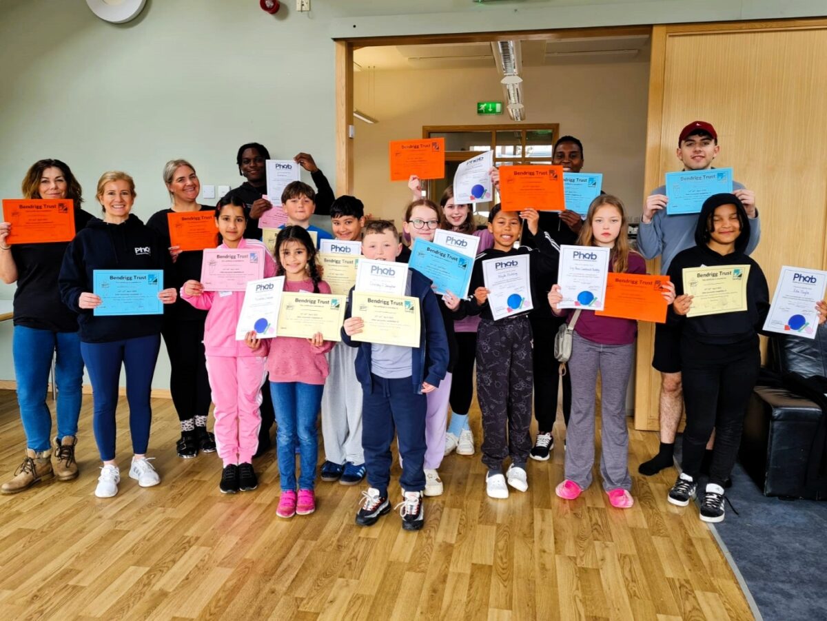 A group of children and adults are standing together smiling holding up different coloured certificates.