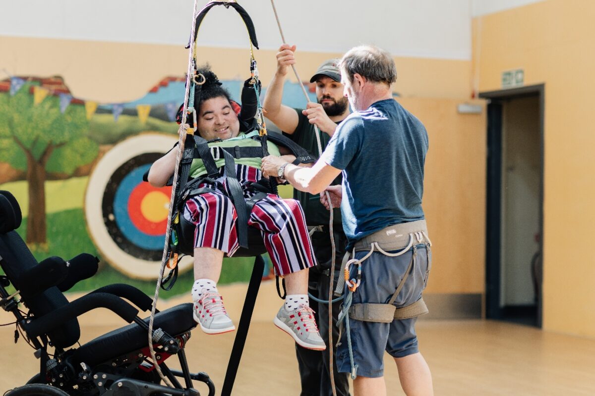 Two people are assisting someone sat in a hoist above the ground. There is a wheelchair beside them.
