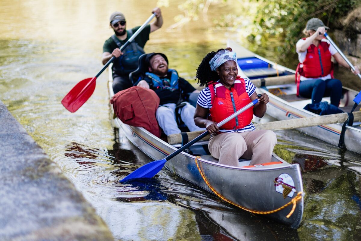 Four people are sat in a metal canoe on a body of water. Three are paddling and smiling, with the person in the middle sat on a beanbag and laughing.