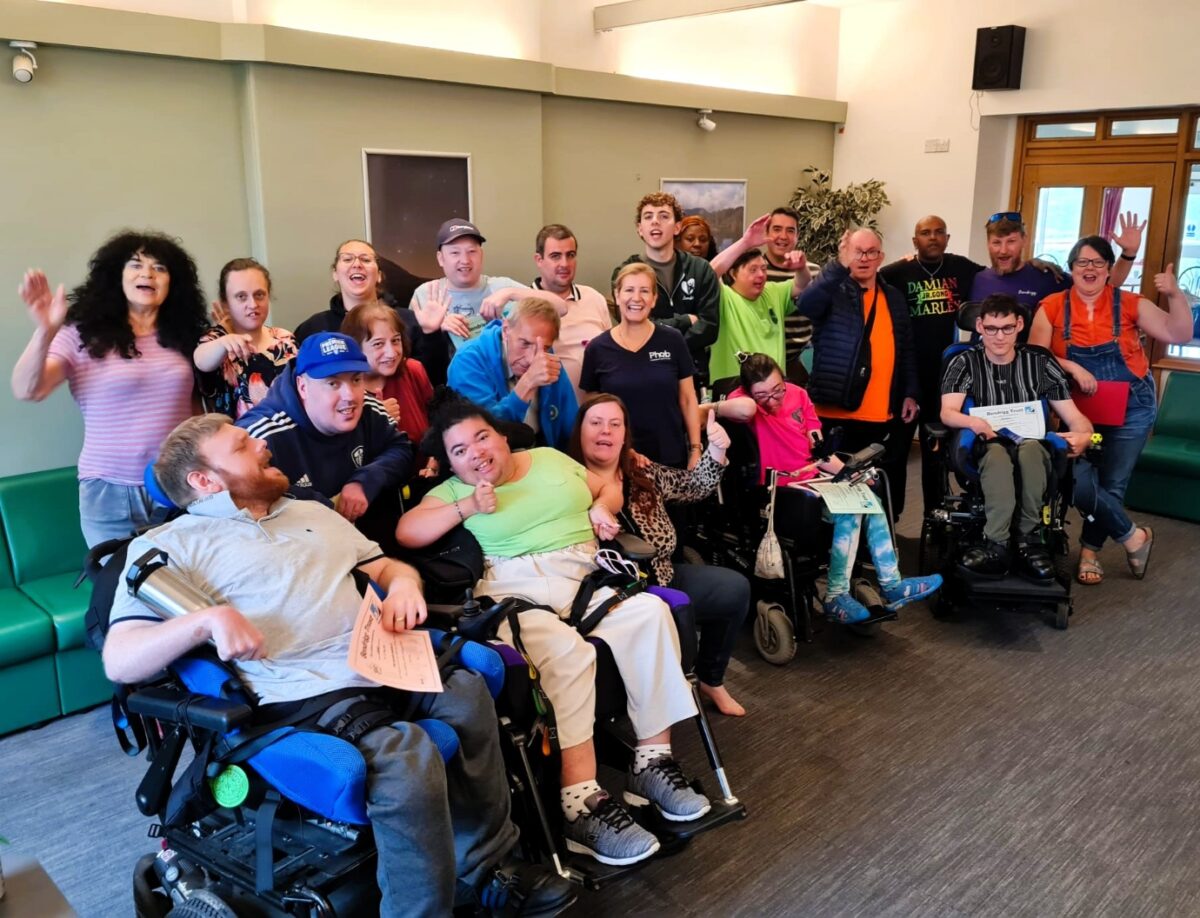 22 people are grouped together inside smiling and waving. Four people are sat in wheelchairs at the front of the group.