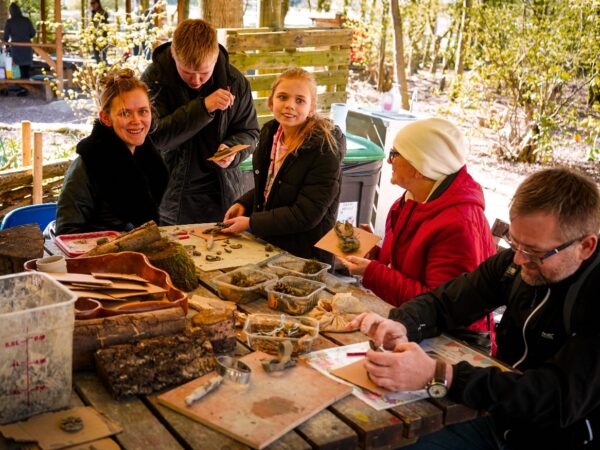 A group of adults and children are sat around a bench using pinecones and other items to make art.