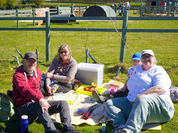 Three adults and a child are sat on a picnic rug in the sunshine next to a fence and smiling.