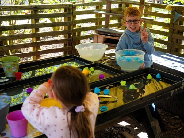 Two children sat outside, with a table with plastic bowls and plastic eggs in front of them. One child is smiling and making a peace sign.