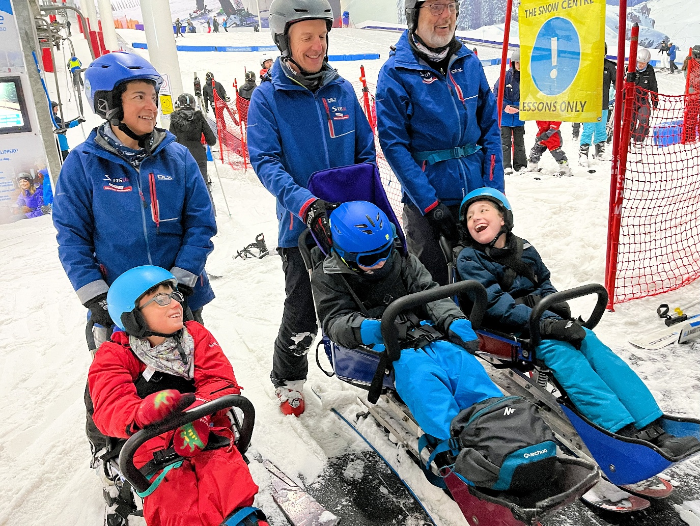 Three children are sat in sit skis, and behind each of them are three adults wearing blue jackets and holding on to the sit ski. Everyone is laughing or smiling.
