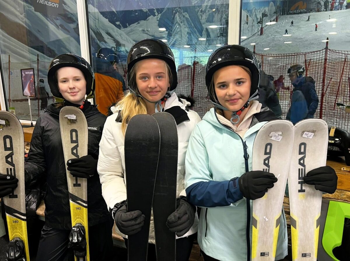 Three children or teenagers are standing together smiling, each holding a set of skis and wearing black helmets. Behind them is an indoor ski hill with snow.