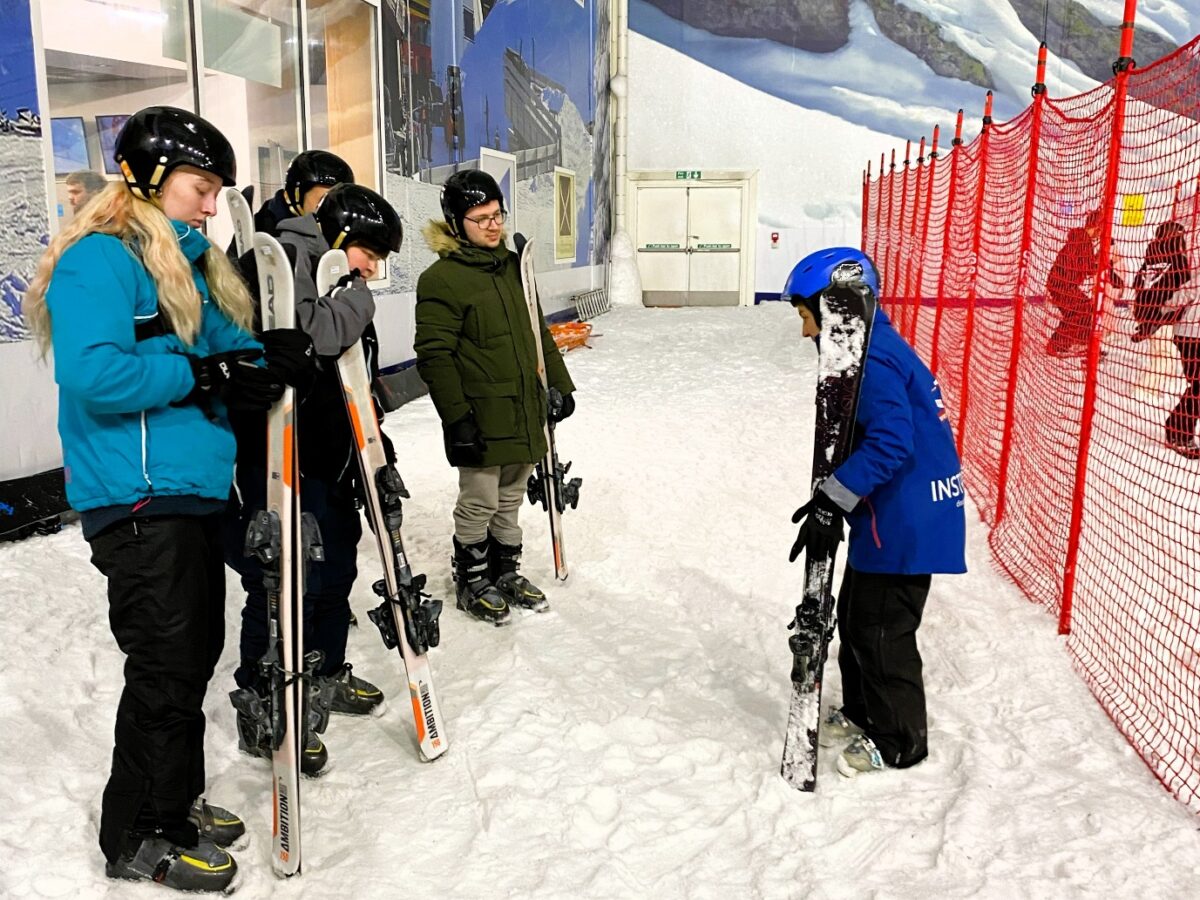 A group of adults are stood in a line wearing black helmets and holding skis, looking towards a person wearing a blue helmet and jacket also holding skis. They are indoors but standing on snow.