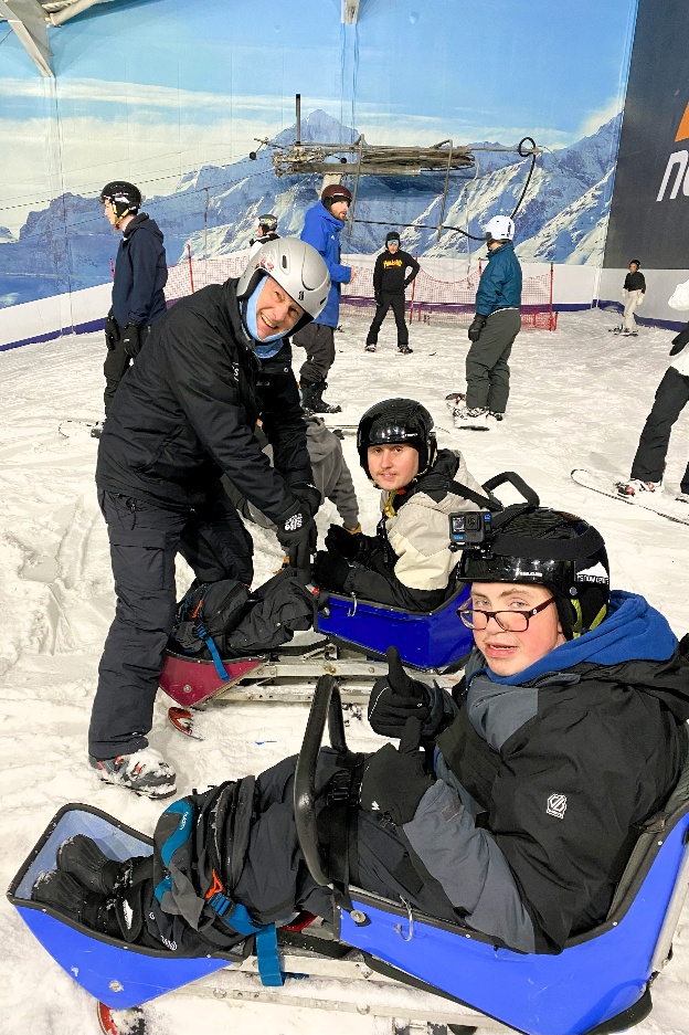 Two young adults are sitting in sit skis, smiling, with an adult holding on to the front of a sit ski, also smiling. You can see snow beneath them and people also wearing skiing gear in the background.
