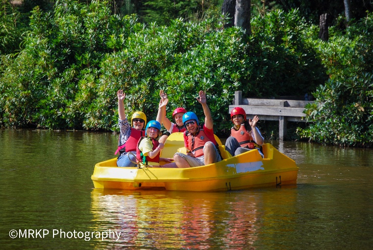 A group of people using a yellow pedalo are waving on a lake.