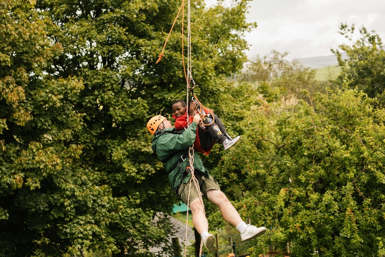 An adult and a child are ziplining together mid-air, smiling. You can see foliage and hills in the background.