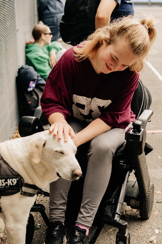 A young adult wearing a red t-shirt is sitting in an electric wheelchair, looking down and stroking a dog.