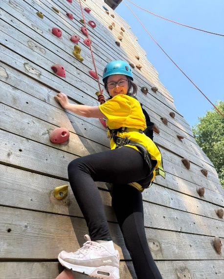 Person wearing a blue helmet and yellow T-shirt climbs an outdoor wooden climbing wall with colourful handholds. The person is secured with a harness and rope, and the sky is clear with trees visible in the background.