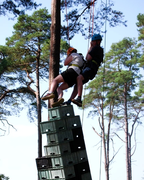 Two people wearing helmets and harnesses are suspended by ropes while balancing on a tall stack of plastic crates outdoors. The crates are stacked vertically, and the scene is surrounded by tall trees under a clear sky.
