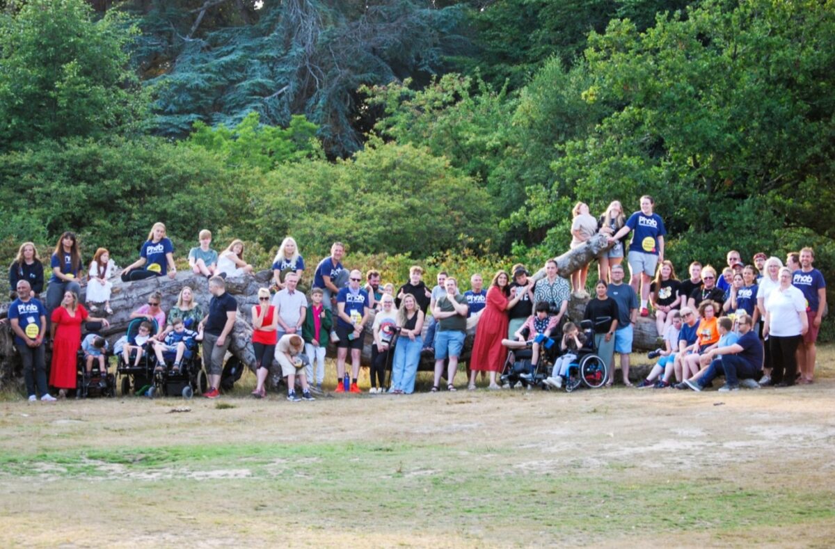 A large group of adults and children is grouped together around a large fallen tree, with some sitting and standing on the tree. People are smiling and waving.
