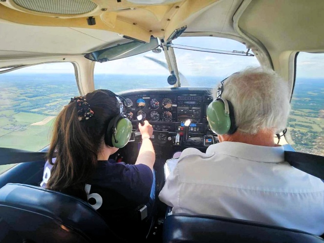 A person wearing a headset sits in the cockpit of a small plane, pointing at the controls while an instructor flies, with countryside visible through the windows below.