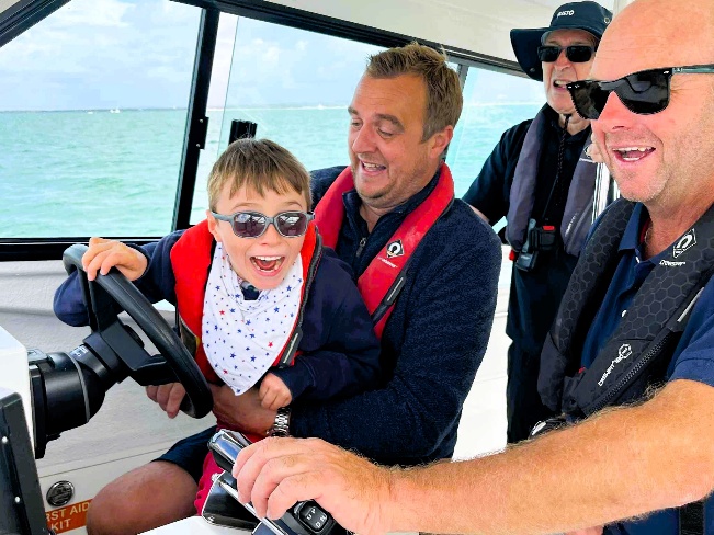 A child steers a boat with help from adults, all wearing life jackets, out on the open sea.