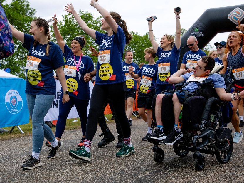 Group of participants wearing event T‑shirts and numbered bibs cheering as they take part in a charity walk, with one person using a wheelchair, outdoors near a finish area.