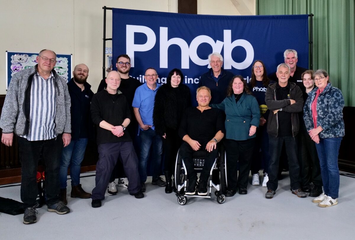 Group of adults standing together for a photo in front of a large Phab banner indoors, with one person seated in a wheelchair at the centre, showing a sense of community and inclusion.