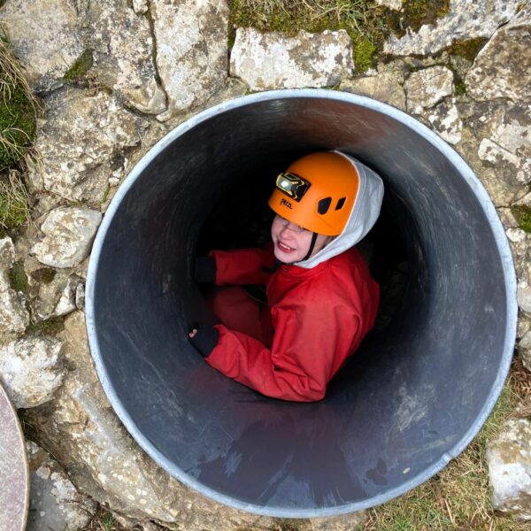 A person wearing an orange helmet and red waterproof suit sits inside a large metal tunnel set into rocky ground, preparing to move through it.
