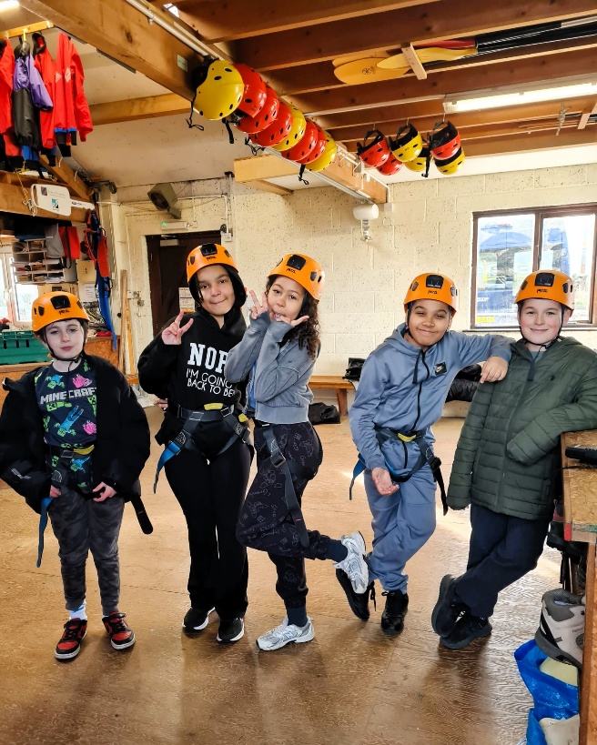 A group of children stand together inside an equipment room, wearing orange climbing helmets and harnesses, smiling and striking fun, energetic poses.
