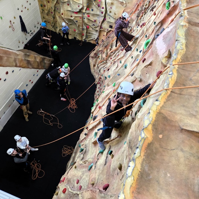 Several people take part in indoor climbing on a tall artificial climbing wall. Some are climbing while others stand below holding ropes for safety.