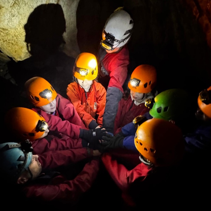 A group of children in orange helmets and red coveralls stand in a dark cave with their hands stacked together in the centre, joined by an adult wearing a white helmet. Their headlamps cast light on their faces and the cave walls, creating deep shadows around them.