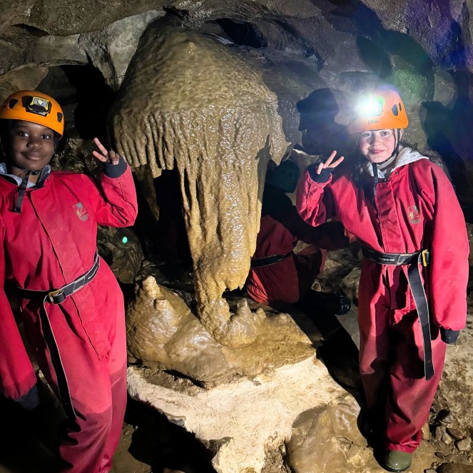 Two children wearing orange helmets with headlamps and red coveralls stand inside a cave beside a large, muddy stalactite formation. Both are smiling and holding up peace signs, with rocky cave walls and formations visible around them.