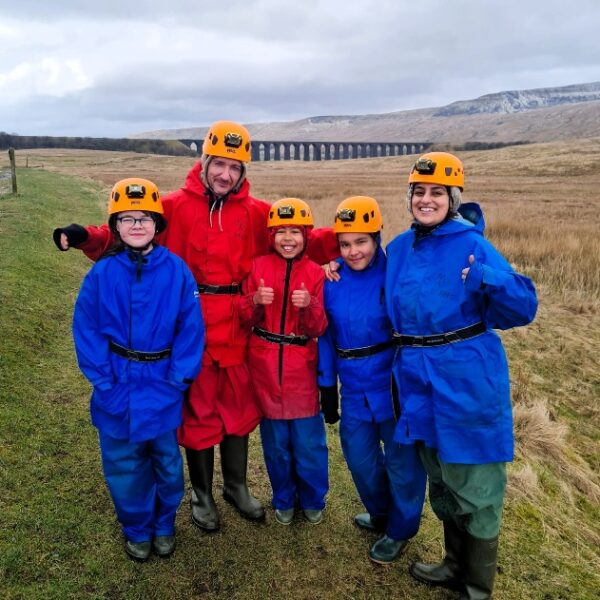 A group of five people wearing helmets, waterproof suits and wellies stand together on grassy countryside. They are dressed in bright red and blue outdoor gear, with rolling hills and a long stone viaduct visible in the background.
