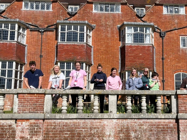 A group of people stand along a stone balcony outside a large red‑brick manor house, enjoying the sunny weather. They are lined up in a row, chatting and looking out over the gardens, with the building’s distinctive bay windows behind them.