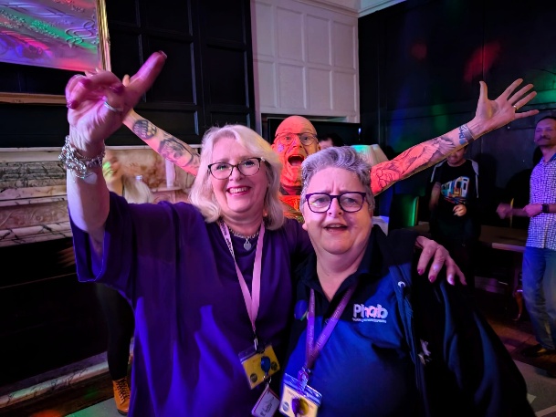 Three people pose together on a lively disco dancefloor, smiling and raising their arms in celebration. Colourful lights illuminate the room, and other people can be seen dancing and enjoying the upbeat atmosphere in the background.