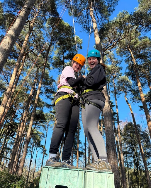 Two people wearing helmets and safety harnesses stand together on top of stacked crates in a forest, taking part in an outdoor adventure activity. Tall pine trees rise around them under a bright blue sky, and ropes from the challenge course stretch overhead.
