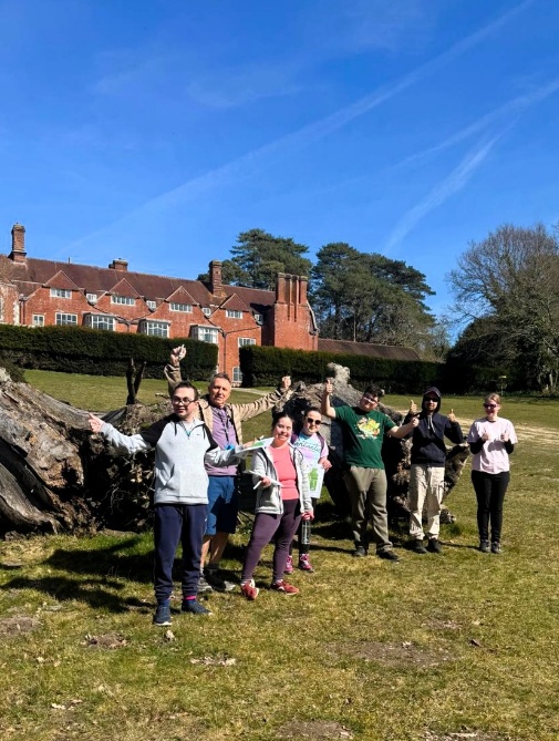 A group of people stand together on a sunny lawn in front of a large red‑brick manor house, posing with their arms raised in celebration. They are gathered beside a large fallen tree trunk, with bright blue sky and tall trees in the background.