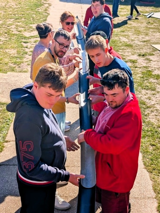 A group of people stand in two lines outdoors, working together to balance and pass a long plastic tube as part of a team‑building activity. They are focused on holding the tube steady, with bright sunlight casting shadows on the ground and grass surround