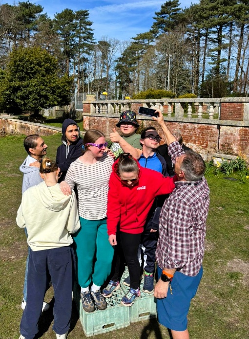 A group of people stand closely together on top of stacked crates outdoors, posing for a selfie being taken by a man holding a phone above them. They are gathered in a sunny garden area with grass, trees, and an old brick wall and balustrade behind them.