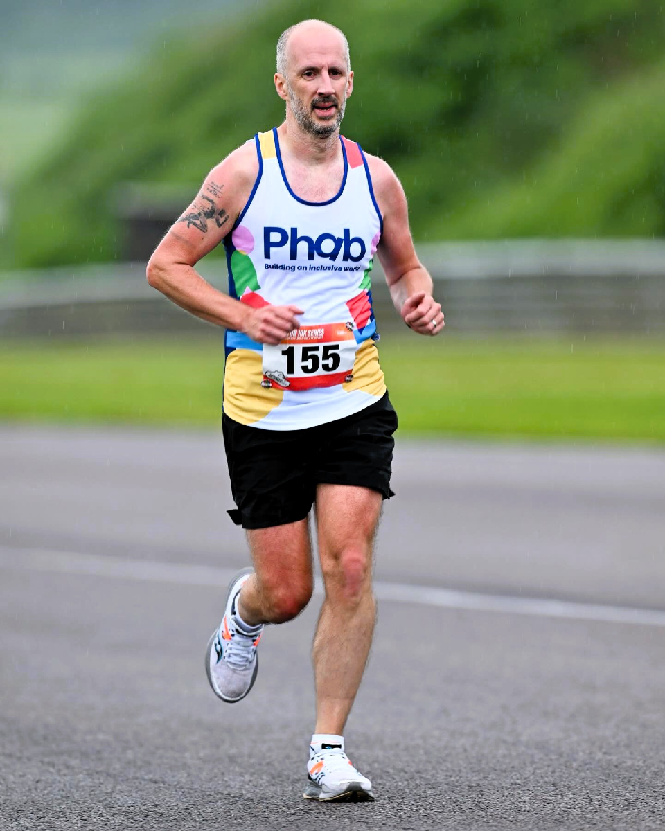 Runner wearing a white “Phab – Building an inclusive world” vest with race number 155 runs along a wet road in light rain, with a blurred green background.