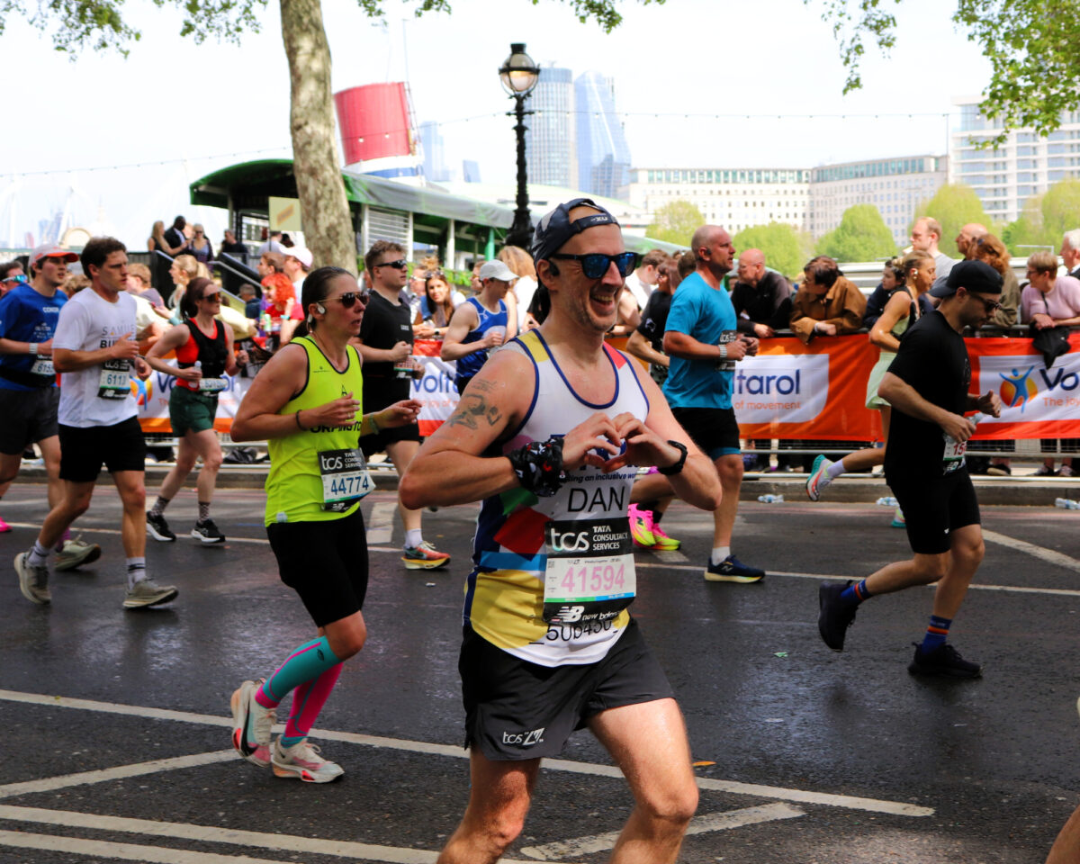 Road race scene with multiple runners moving past barriers lined with spectators and banners. One runner in the foreground wearing a “Phab” vest with the name “Dan” and a race bib number forms a heart shape with their hands while running.
