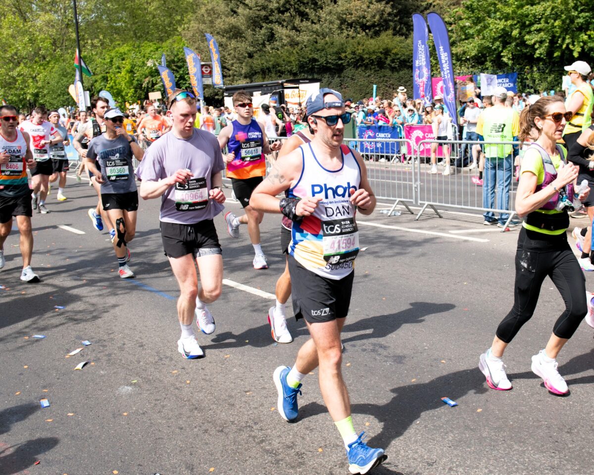Another road race moment showing several runners moving along a closed city street. A runner in a “Phab” vest with the name “Dan” and a race bib is among the group, with cheering spectators, flags, and charity banners lining the route.
